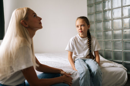 Portrait of blonde young adult mother and cute child enjoy talking relaxing sitting on bed in light...