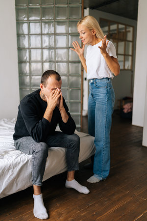 Vertical shot of frustrated Caucasian husband sitting on bed ignoring angry standing blonde wife arguing blaming upset man of problems at home. Jealous woman shouting at sad boyfriend.の写真素材