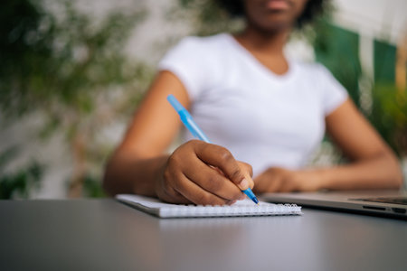 Close-up cropped shot of unrecognizable African-American woman holding pen writing working from home, noting writing down, housewife planning week write to-do list, sitting at table with laptop.の写真素材