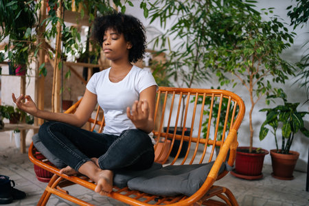 Serene calm black young woman sitting with closed eyes on sofa in lotus pose, practicing yoga meditation at home, relaxing and breathing deep for mental health in room with biophilia design.の写真素材