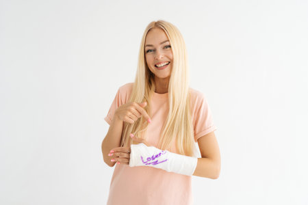 Studio portrait of smiling blonde young woman with broken arm wrapped in plaster bandage with funny...