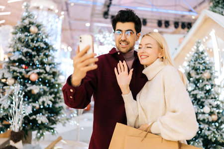Portrait of friendly young couple talking looking at smartphone having video call, giving online New Year greetings standing in hall of celebrate shopping mall with xmas decorations in Christmas eveの写真素材