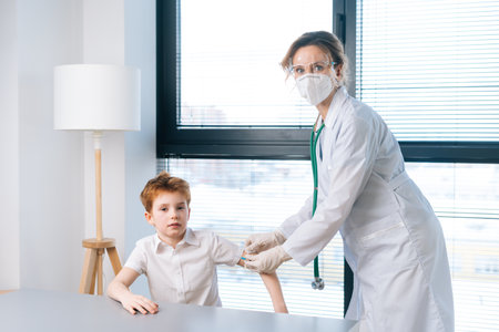 Portrait of female doctor applying plaster on shoulder of child boy after vaccination injection at hospital by window, looking at camera. Concept of vaccination program, prevention of diseases.の写真素材