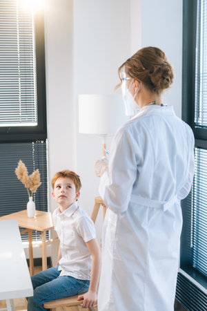 Vertical shot of unrecognizable female doctor in latex gloves preparing vaccine for child boy at hospital with light interior. Concept of vaccination program, prevention of infectious diseases.の写真素材