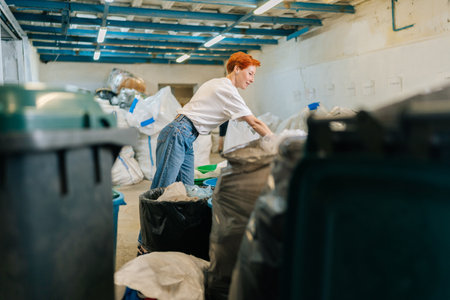 Wide shot of focused female volunteer worker in latex gloves sorting diverse waste for further disposal at modern private waste recycling plant. Concept of waste separation and sustainable lifestyle.の写真素材