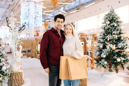 Portrait of elegant young couple enjoying shopping together, holding bags with gifts and looking at camera standing hugging in hall of celebrate shopping mall, with xmas decorations in Christmas eve