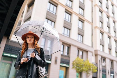 Low-angle view of charming redhead young woman in fashion hat waiting in city street holding transparent umbrella in rainy weather rain, looking away. Concept of female lifestyle at autumn season.の写真素材
