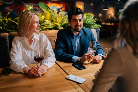 Elegant bearded man in suit and two pretty blonde women sitting restaurant talking laughing making toasts having fun, celebrating birthday. Group of happy friends drinking and toasting wine in bar.の写真素材