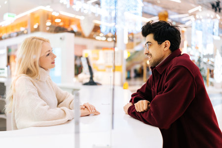 Side view of happy young couple of elegant bearded man in glasses and pretty blonde woman chatting through glass partition standing in hall of shopping mall. Concept of lifestyle social distancing.の写真素材