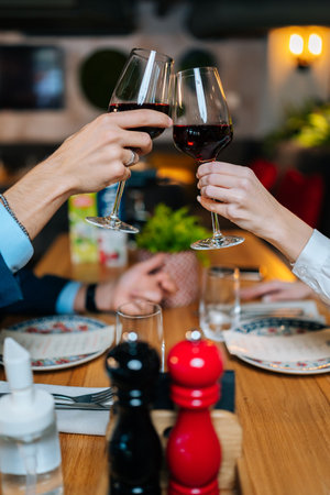 Close-up vertical shot of unrecognizable couple clinking glasses with red wine sitting at table in restaurant at evening. Happy man and woman enjoying nice romantic dinner, celebrating anniversary.の写真素材