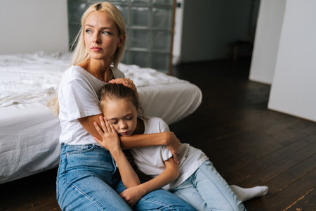 Portrait of upset cute little girl and loving caring mother looking away comforting offended afraid child daughter, showing love and care, expressing support, hugging, stroking hair sitting on floor.の写真素材