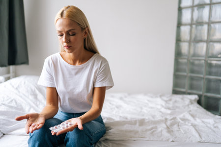 Portrait of disappointed sad pretty young woman holding pills in hands and thinking about taking drugs sitting on soft bed in light room at home. Concept of healthy lifestyle.の写真素材