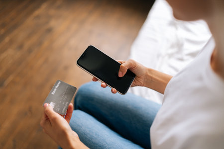 Close-up cropped shot of unrecognizable young woman buying online with credit card using mobile phone, doing online transactions at home. High-angle view of woman shopper entering credit card number.の写真素材