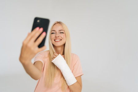 Studio portrait of joyful female blogger with broken arm wrapped in plaster bandage talking smartphone making video call, taking selfie picture, standing on white isolated background.の写真素材