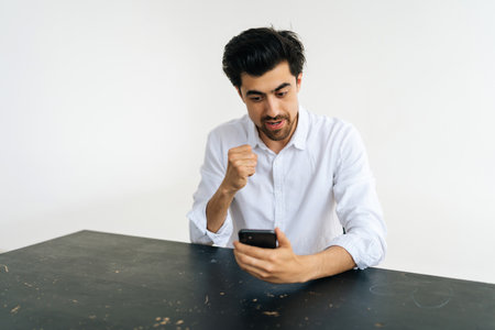 Studio portrait of emotional young bearded man in casual shirt celebrating success, looking at...