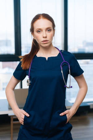 Vertical portrait of confident pretty female doctor wearing medical uniform looking at camera standing on background of window. Woman surgeon posing with stethoscope at hospital room.の写真素材