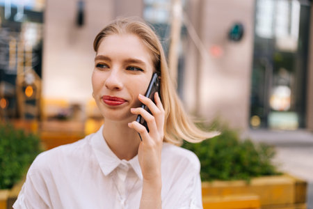 Close-up portrait of happy charming young blonde woman talking on smartphone sitting on bench on city street, smiling looking away. Beautiful lovely lady walking outdoors, talking on mobile phone.の写真素材