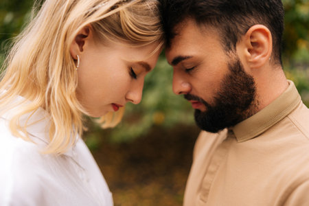 Close-up of loving married couple standing foreheads touched with closed eyes. Happy young bearded man and pretty blonde woman on romantic date in summer park. Couple relaxing heads embracing.の写真素材