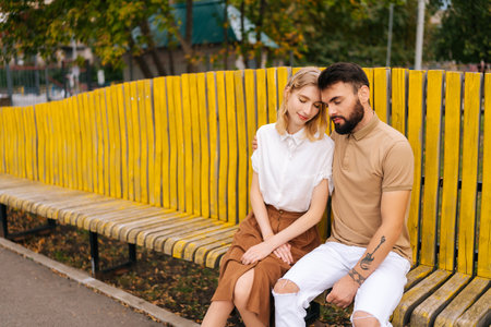 Happy young couple of tourists resting in city park sitting hugging with closed eyes on bench in summer day. Handsome bearded man and attractive blonde woman enjoying spending time togetherの写真素材