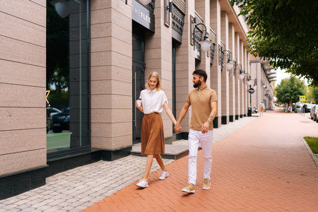 Full length portrait of happy young couple in love walking holding hands on city street. Stylish bearded man and blonde woman holding cup with takeaway coffee enjoying leisure time together outdoors.の写真素材