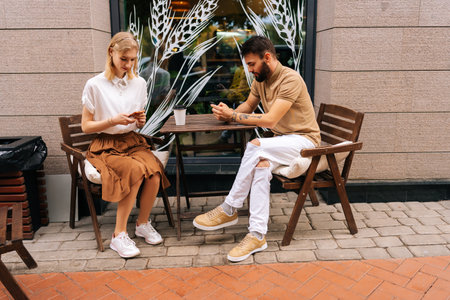 Side view of young couple using looking smartphones, ignoring each other sitting at table with coffee in street cafe on summer day. Concept of internet more interesting than real communication.の写真素材