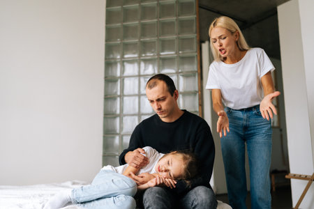 Depressed caring young father embracing loving little daughter sitting on bed while aggressive mother angrily shouting at them in living room. Angry wife scolding, raising voice, scream at family.の写真素材