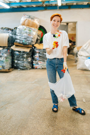 Vertical full length portrait of smiling woman volunteer holding in hands used plastic objects and looking at camera in private waste recycling plant. Concept of environment protection and zero waste.の写真素材