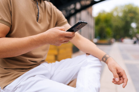 Cropped shot of unrecognizable tattooed male sitting on bench and using typing smartphone looking to screen device on city street. Unknow man in fashion clothes reading message on phone on summer day.の写真素材