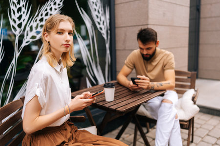 Pretty blonde woman looking at camera, bearded tattooed man using smartphones sitting at table with coffee in street cafe on summer day. Concept of internet more interesting than real communication.の写真素材