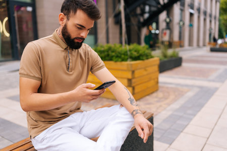 Side view of confident bearded male sitting on bench and using typing mobile looking to screen device on city street. Bearded handsome male in casual clothes reading message on phone on summer day.の写真素材