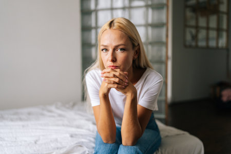 Close-up portrait of pensive young blonde woman sitting alone at in living room and sad looking camera holding hands on chin, thinking over health problems, feeling sadness, boredom, apathy.の写真素材