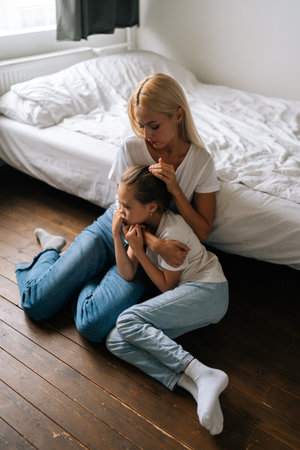 Vertical portrait of loving young mother comforting offended sad little child daughter, showing love and care, expressing support, hugging and stroking hair sitting on floor. Family bonding concept.の写真素材
