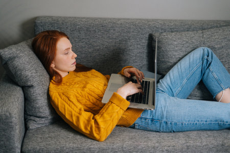 Side view of tired young woman typing on laptop while lying on back on sofa at home. Exhausted redhead female freelancer working chatting in internet by computer lying on couch in living room.の写真素材