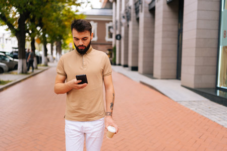 Portrait of confident handsome young man with tattooed hand walking on city street, using smartphone and drinking takeaway coffee on summer day. Bearded hipster male typing mobile phone.の写真素材