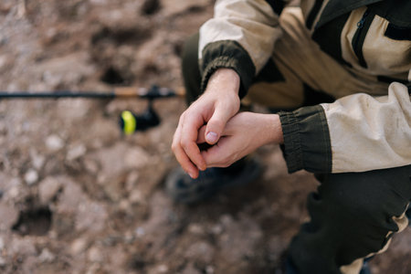 Close-up high-angle view hands of unrecognizable fisherman wearing raincoat sitting on river bank on travel chair with fishing rod waiting for catch on summer day. Concept of leisure activity outdoorsの写真素材