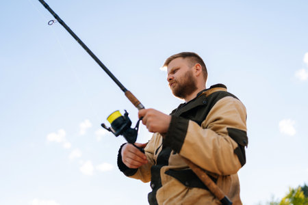 Close-up low-angle view of bearded fisherman holding casting rod wearing raincoat standing on bank waiting for bites on water river at summer day. Concept of lifestyle, leisure activity on nature.の写真素材