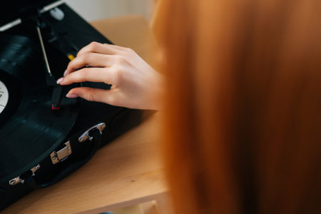 Close-up high-angle view from back of unrecognizable redhead female listening to retro music from vinyl record player. Pretty lady enjoying home entertainment, using retro media technology.の写真素材