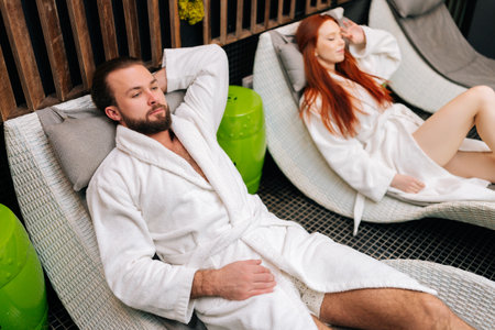 High-angle view of young couple in white bathrobes lying on comfy loungers by poolside after spa treatment at beauty salon. Bearded man and pretty woman enjoying relaxing after body massage treatmentの写真素材