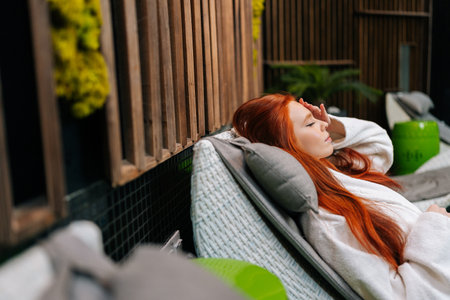 Closeup side view of tranquil young woman in white bathrobe relaxing lying on deck chair with eyes closed by poolside after beauty treatment at spa salon. Calm female enjoying tranquility on vacationsの写真素材