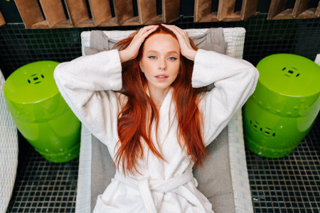 High-angle portrait of charming young woman in white bathrobe relaxing lying on deck chair after beauty treatment at spa salon, looking at camera. Pretty redhead female enjoying wellness weekend aloneの写真素材