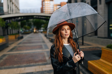 Portrait of elegant young woman in fashion hat standing on beautiful city street with transparent umbrella enjoying rainy weather outdoors, smiling looking at camera. Concept of female lifestyle.の写真素材