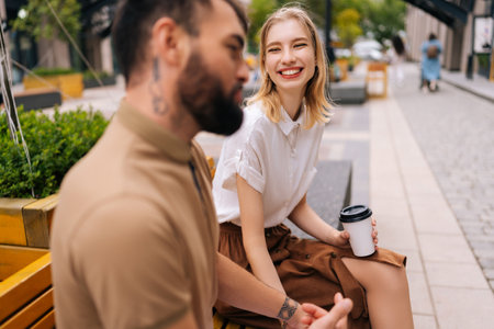 Selective focus view of cheerful young woman having fun communication with bearded tattooed man sitting on bench in city park. Loving couple enjoying pleasant conversation outdoors.の写真素材