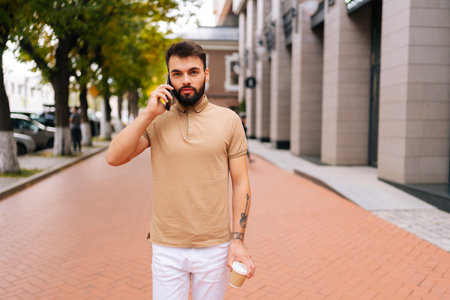 Portrait of bearded handsome young man with tattooed hand walking on city street, talking on smartphone and drinking takeaway coffee on summer day. Confident male having conversation speaking on phoneの写真素材
