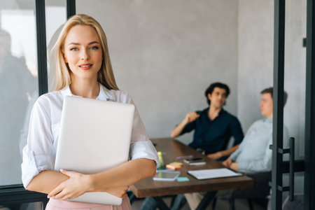 Portrait of successful startup businesswoman in casual clothes holding laptop in hand standing in office, looking at camera. Young business team discussing project sitting at desk on background.の写真素材