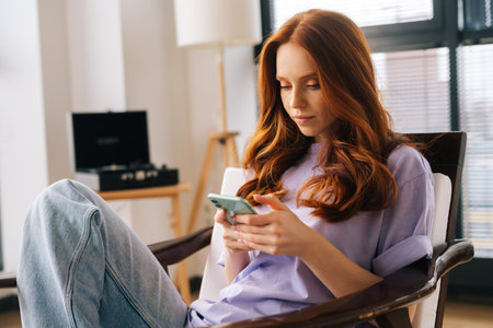 Portrait of thoughtful focused young woman texting message on mobile phone sitting on arm chair. Pensive female using cellphone at home on background of window. Pretty lady chatting online on cell.の写真素材