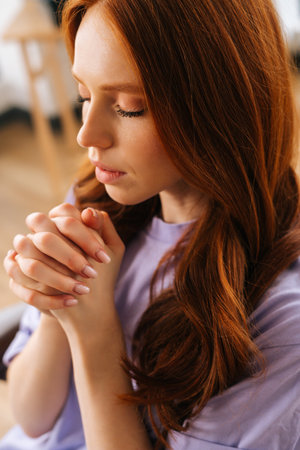 Vertical closeup portrait of sad young woman sincerely praying with eyes closed holding folded hands in front of face at home. Worried female put hands in prayer praying excitedly with hope.の写真素材