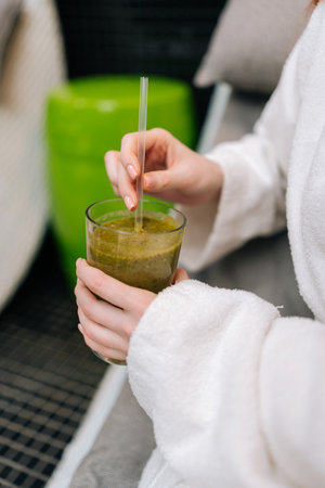 Vertical closeup cropped shot of unrecognizable young woman in white bathrobe holding glass with delicious healthy freshly squeezed vitamin juice in hand, sitting on lounger at spa salon.の写真素材