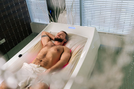 High-angle view of handsome bearded young man lying relaxing on electric massage bed and enjoying spa treatment at luxury spa salon. Muscular male having rest and enjoying wellness weekend alone.の写真素材