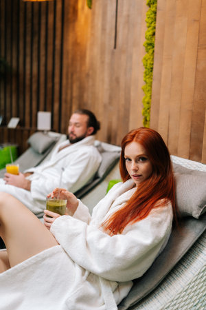 Vertical portrait of pretty young woman and bearded man in white bathrobes lying on loungers by poolside, drinking fresh fruit cocktail at spa salon, after massage treatment, looking at camera.の写真素材