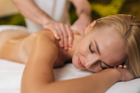 Close-up face of relaxed young woman with closed eyes getting professional back massage in spa salon. Beautiful naked female client with perfect skin having massage. Concept of healthy lifestyleの写真素材